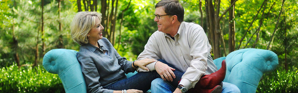 Barbara and Dennis Rainey looking at each other and smiling while sitting on a couch in a wooded area.